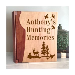 May include: A wooden photo album with a light brown wood cover and a dark brown wood spine. The cover is engraved with the text "Anthony's Hunting Memories" and features a deer, a fawn, trees, and birds.