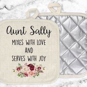 May include: A set of two beige pot holders with a floral design. One pot holder has the text "Aunt Sally mixes with love and serves with joy".
