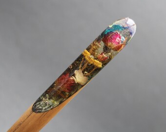 Puede incluir: Un palo de pelo de madera con una parte superior de resina transparente que contiene flores prensadas y purpurina iridiscente. Las flores son rosas, amarillas y blancas.