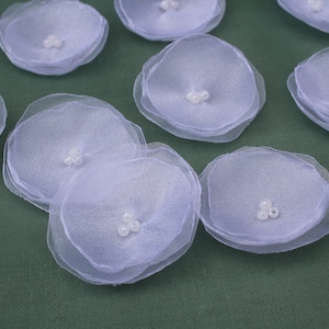 May include: A collection of eight white, layered fabric flowers with pearl accents. The flowers are arranged on a green surface.