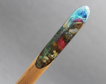 Puede incluir: Un taco de madera con una punta de resina transparente llena de purpurina de colores y flores secas. La resina es de un azul, verde y rojo vibrantes con purpurina dorada.