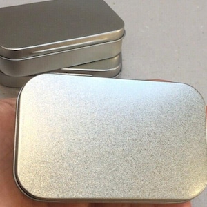 May include: A silver metal rectangular tin with rounded corners and a lid, held in a hand. Several similar tins are stacked in the background. The tins have a brushed metal finish. The tins are made of metal.