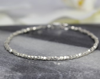 May include: A delicate silver-toned bracelet composed of numerous small, faceted, irregular metallic beads strung together in a continuous circular chain. The surface of each bead reflects light, creating a textured appearance along the entire length.