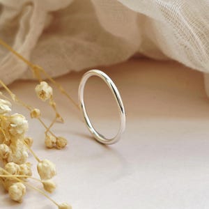 May include: A simple, polished silver band ring with a smooth, rounded surface. The ring is positioned upright, showcasing its clean, circular shape and reflective metallic finish, standing out against a soft, neutral-toned surface.