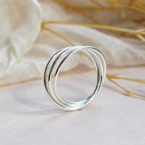 May include: A polished silver ring featuring a multi-band design composed of three thin, interconnected circular wires that overlap to create a layered appearance. The metal surface is smooth and reflective, showcasing a bright, metallic finish.