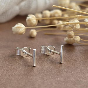 May include: A pair of silver stud earrings featuring a simple horizontal bar design. Each earring consists of a thin, polished metal bar attached to a post with a matching silver butterfly clutch backing, showcasing a clean and metallic finish.