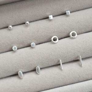 May include: A collection of six pairs of silver stud earrings arranged in rows. The set includes small spheres, squares, open circles, flat discs, ovals, and thin vertical bars. Each earring features a polished metallic finish and a simple geometric shape.