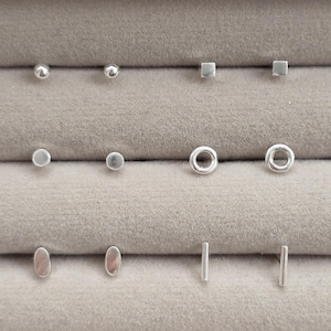 May include: A collection of six pairs of silver-toned stud earrings arranged in three rows. The set includes small spheres, tiny squares, flat circles, open rings, oval shapes, and thin vertical bars, all featuring a polished metallic finish.