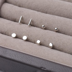 May include: A selection of silver stud earrings in different shapes: bars, spheres, circles, and squares. The earrings are presented in a grey jewellery box, highlighting their minimalist design and polished finish. These small, delicate earrings are suitable for everyday wear.