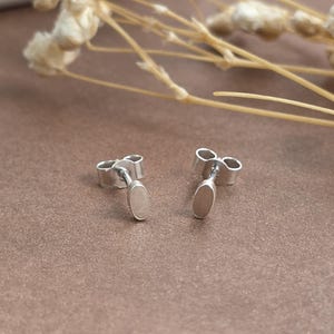 May include: A pair of silver stud earrings featuring a small, smooth, vertical oval-shaped front piece attached to a thin post with a standard butterfly-style earring back closure. The metal has a polished, reflective finish.