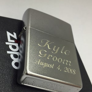 May include: A silver Zippo lighter with engraved text reading "Kyle Groom August 4, 2018." The lighter is resting on a black box with the Zippo logo. The lighter has a brushed metal finish.
