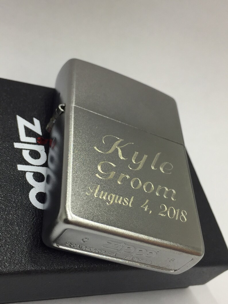 May include: A silver Zippo lighter with engraved text reading "Kyle Groom August 4, 2018." The lighter is resting on a black box with the Zippo logo. The lighter has a brushed metal finish.