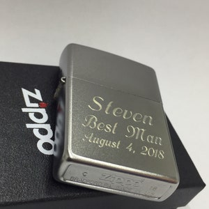 May include: A silver Zippo lighter engraved with "Steven Best Man August 4, 2018." The lighter is on top of a black box with the Zippo logo. The lighter is made in Bradford, PA.