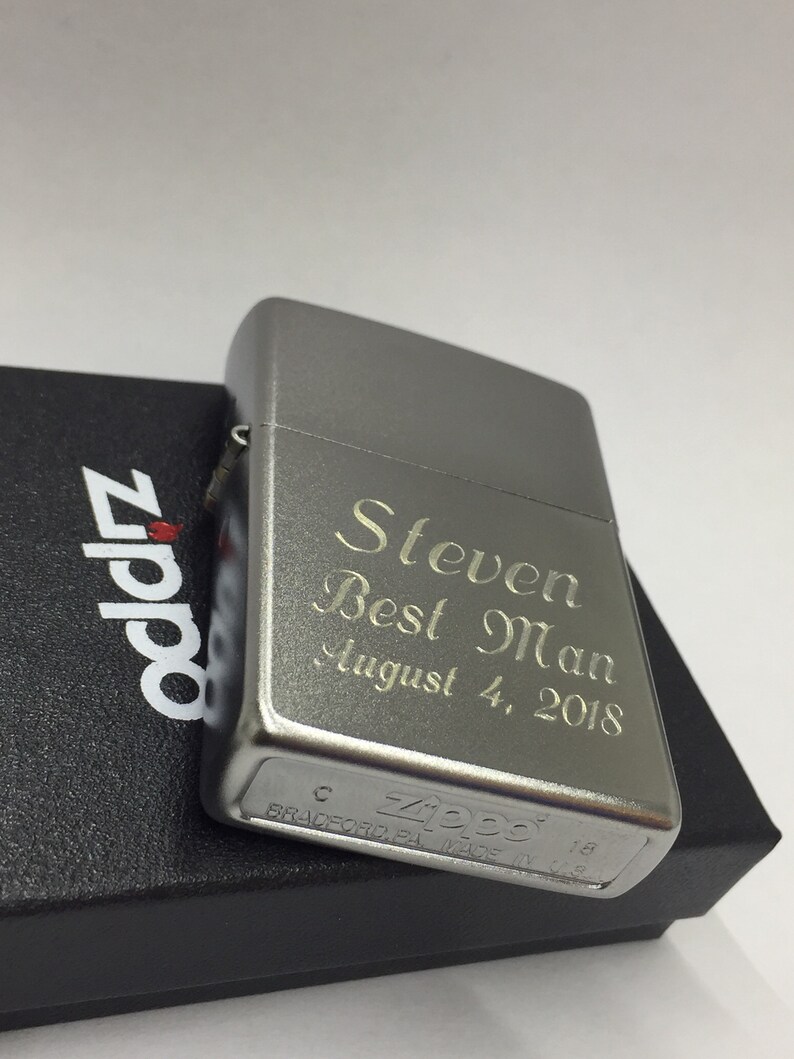 May include: A silver Zippo lighter engraved with "Steven Best Man August 4, 2018." The lighter is on top of a black box with the Zippo logo. The lighter is made in Bradford, PA.