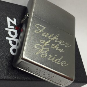 May include: A gray metal Zippo lighter with the inscription "Father of the Bride" in a cursive font. The lighter is resting on a black box with the Zippo logo. A classic gift for a wedding.