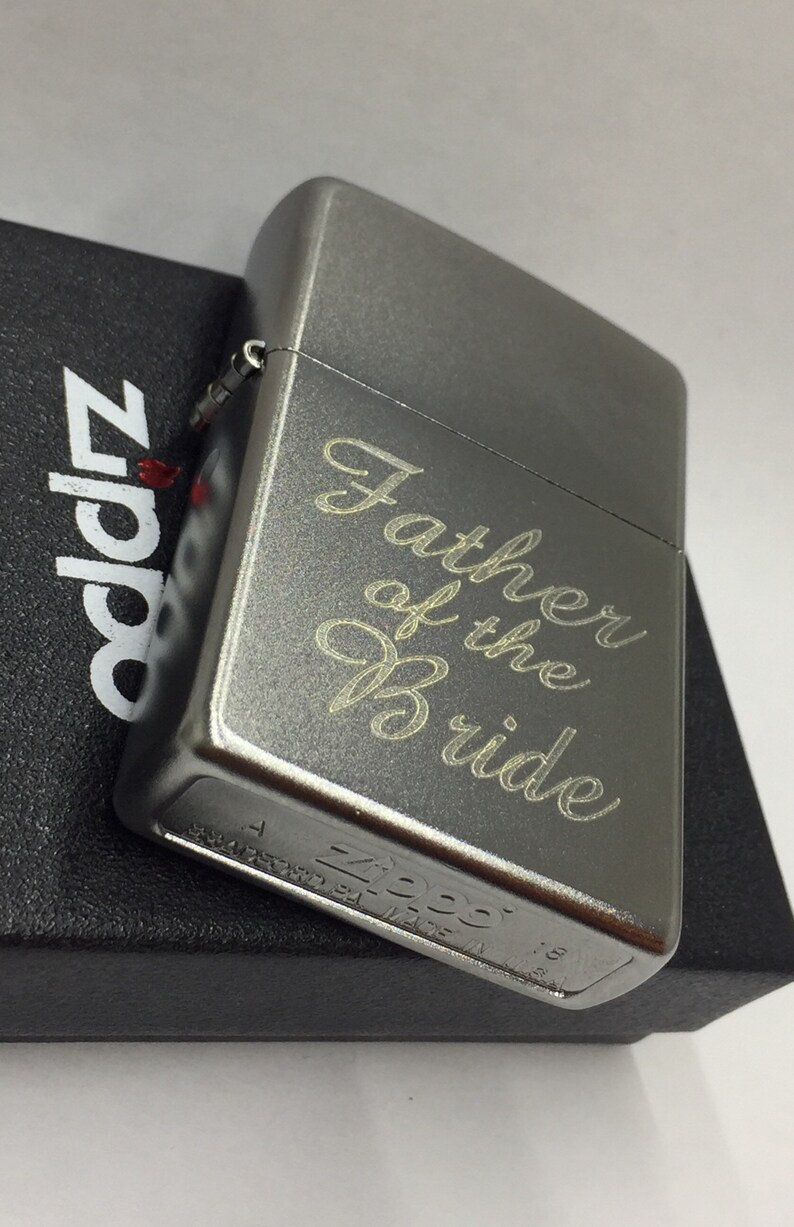 May include: A gray metal Zippo lighter with the inscription "Father of the Bride" in a cursive font. The lighter is resting on a black box with the Zippo logo. A classic gift for a wedding.
