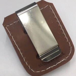 May include: A brown leather money clip with a shiny silver metal clip. The leather is stitched with white thread around the edges. The text "MADE IN U.S.A." is visible on the leather.