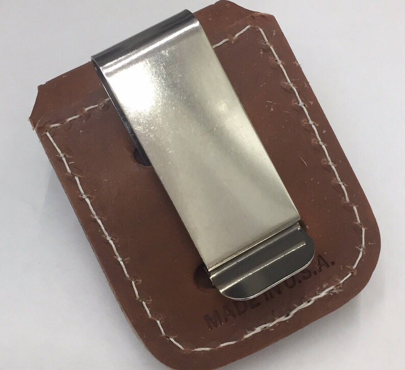 May include: A brown leather money clip with a shiny silver metal clip. The leather is stitched with white thread around the edges. The text "MADE IN U.S.A." is visible on the leather.
