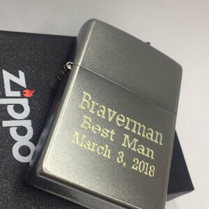 May include: A gray metal lighter with the text "Braverman Best Man March 3, 2018" engraved in gold. The lighter is resting on a black box with the Zippo logo.