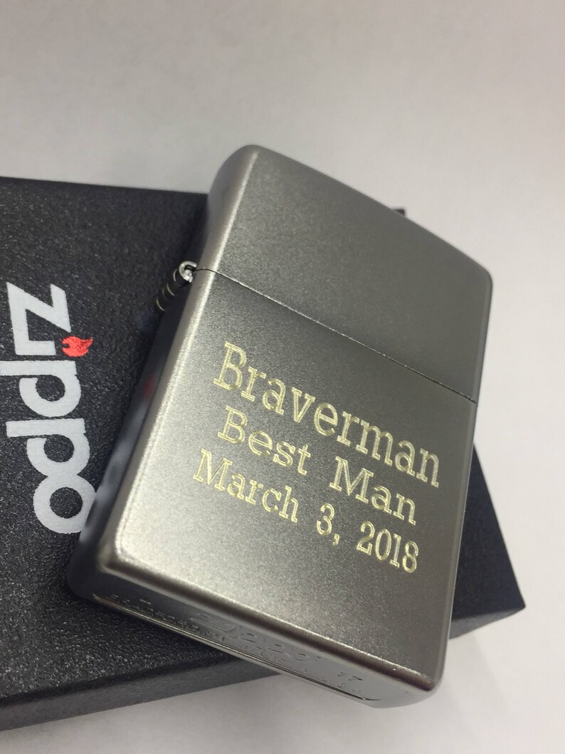 May include: A gray metal lighter with the text "Braverman Best Man March 3, 2018" engraved in gold. The lighter is resting on a black box with the Zippo logo.