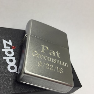May include: A silver Zippo lighter with the engraved text "Pat Groomsman 9/22/18." The lighter is resting on a black textured box with the Zippo logo in red and white.