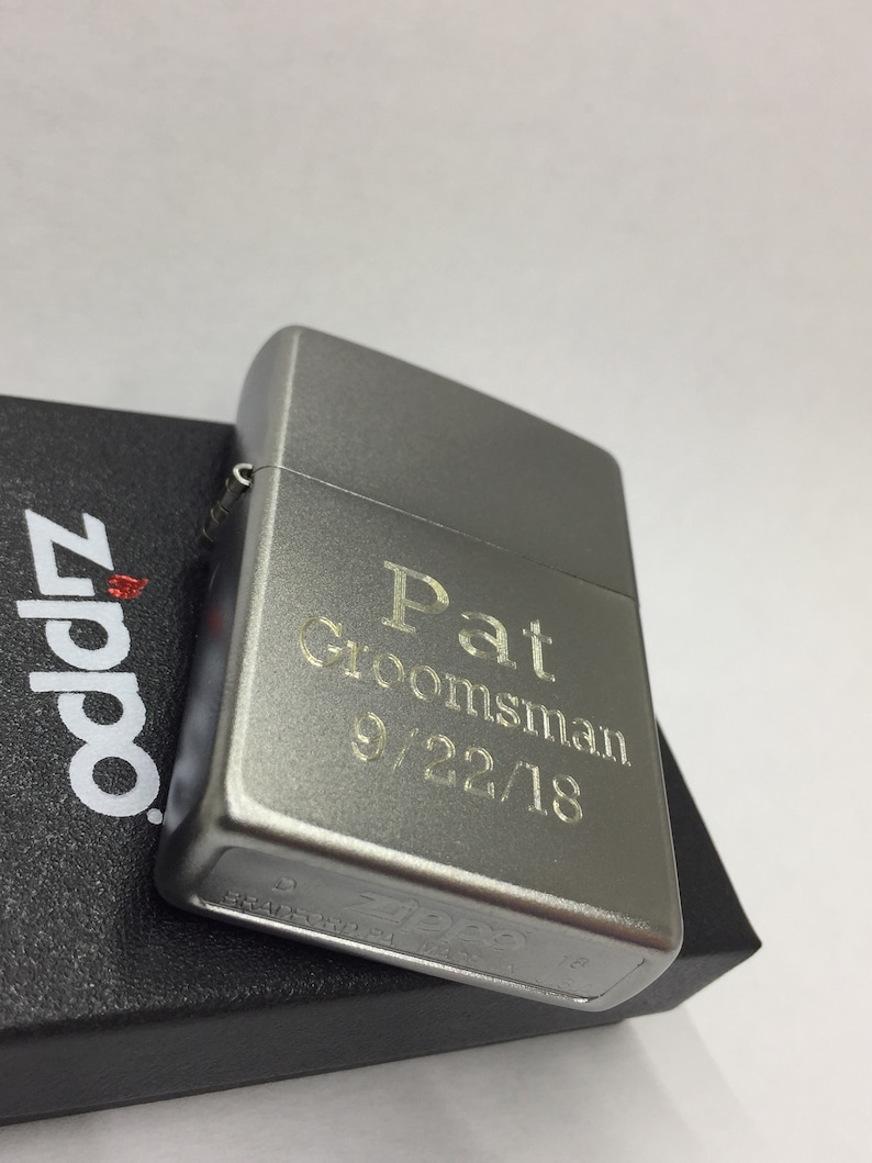 May include: A silver Zippo lighter with the engraved text "Pat Groomsman 9/22/18." The lighter is resting on a black textured box with the Zippo logo in red and white.