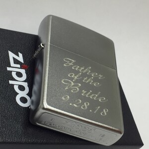 May include: A silver Zippo lighter engraved with "Father of the Bride 9.28.18". The lighter is resting on a black box with the Zippo logo. The lighter has a brushed metal finish and a small hinge.