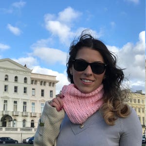 May include: A woman wearing a pink crocheted infinity scarf and sunglasses, standing in front of a white building with a blue sky in the background.