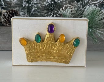 May include: A gold-colored crown with colorful jewels, mounted on a white canvas. The crown features a textured surface and is adorned with purple, green, and yellow gemstones. The canvas has a gold border.