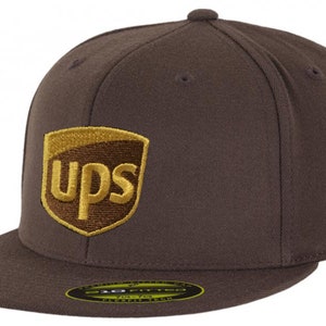 Puede incluir: Gorra de béisbol marrón con visera plana. La gorra presenta el logotipo de UPS en dorado y marrón en la parte delantera. El sombrero es de estilo Flexfit.
