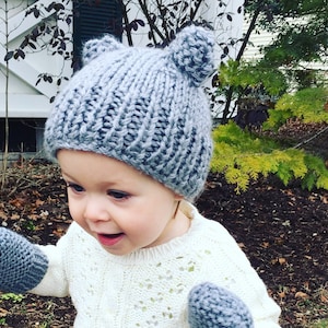 May include: A toddler wearing a gray knitted hat with ear flaps and matching mittens. The hat and mittens are made with a chunky yarn and have a simple knit stitch.
