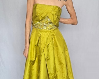 May include: A strapless, chartreuse-colored dress with a fitted bodice and a flared skirt. The bodice features a decorative bow and a sequined band at the waist. The dress is made of a shiny fabric, and the overall style is elegant and formal.