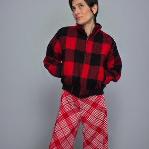 A woman wearing a red and black plaid jacket and red and white plaid pants stands against a gray background.