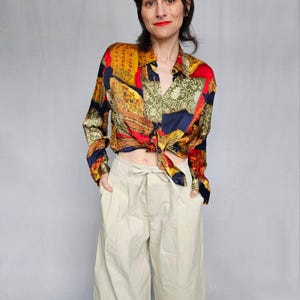 May include: A woman wearing a multi-coloured silk shirt with a paisley pattern. The shirt is tied at the waist and the woman is wearing beige wide-leg trousers.
