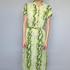 May include: A short-sleeved dress with a snake print in shades of green and black. The dress has a collared neckline, a button-down front, and a matching belt. The dress falls to the mid-calf.