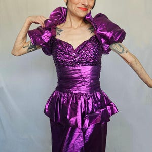 May include: A striking, metallic purple dress with a sweetheart neckline and a peplum waist. The dress features large, ruffled shoulder accents and a fitted bodice. The fabric has a shiny, reflective quality, creating a bold and eye-catching look.