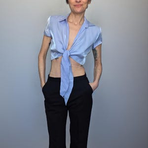 A woman wearing a blue tie-front blouse and black pants stands against a gray background, looking to the side with a neutral expression.