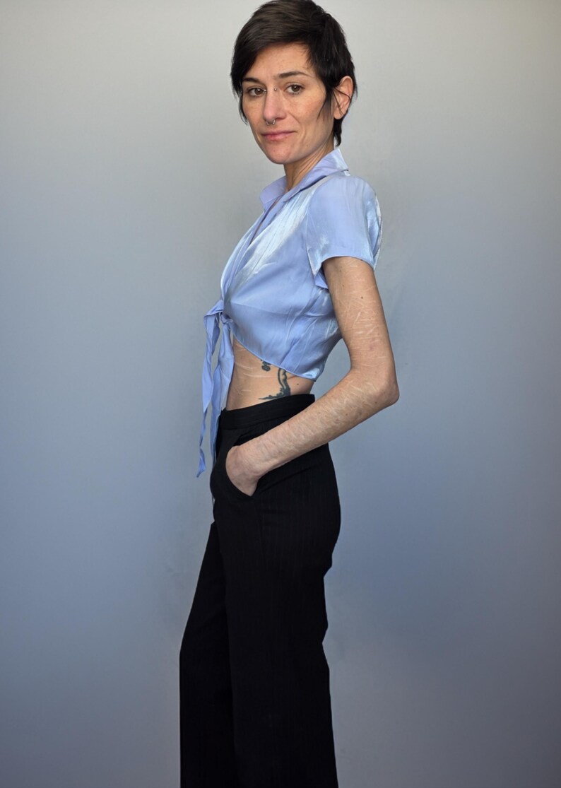 A woman wearing a blue blouse and black pants stands against a gray background, looking directly at the camera with a neutral expression.