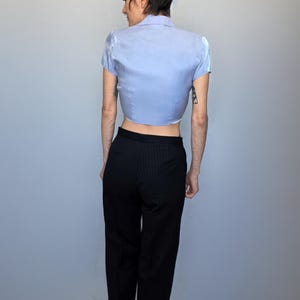 A person wearing a blue shirt and black pants is standing against a plain gray background.