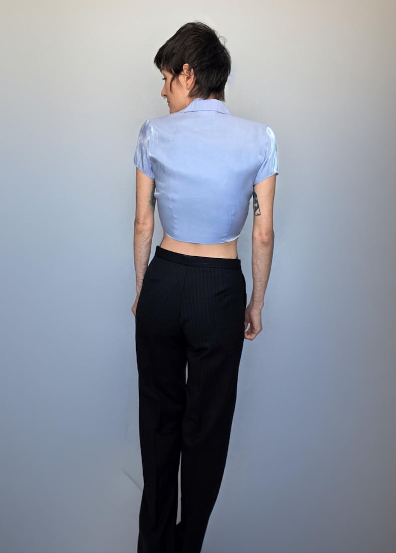 A person wearing a blue shirt and black pants is standing against a plain gray background.