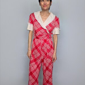 A woman wearing a red and white checkered jumpsuit with a lace collar and short sleeves stands against a gray background.