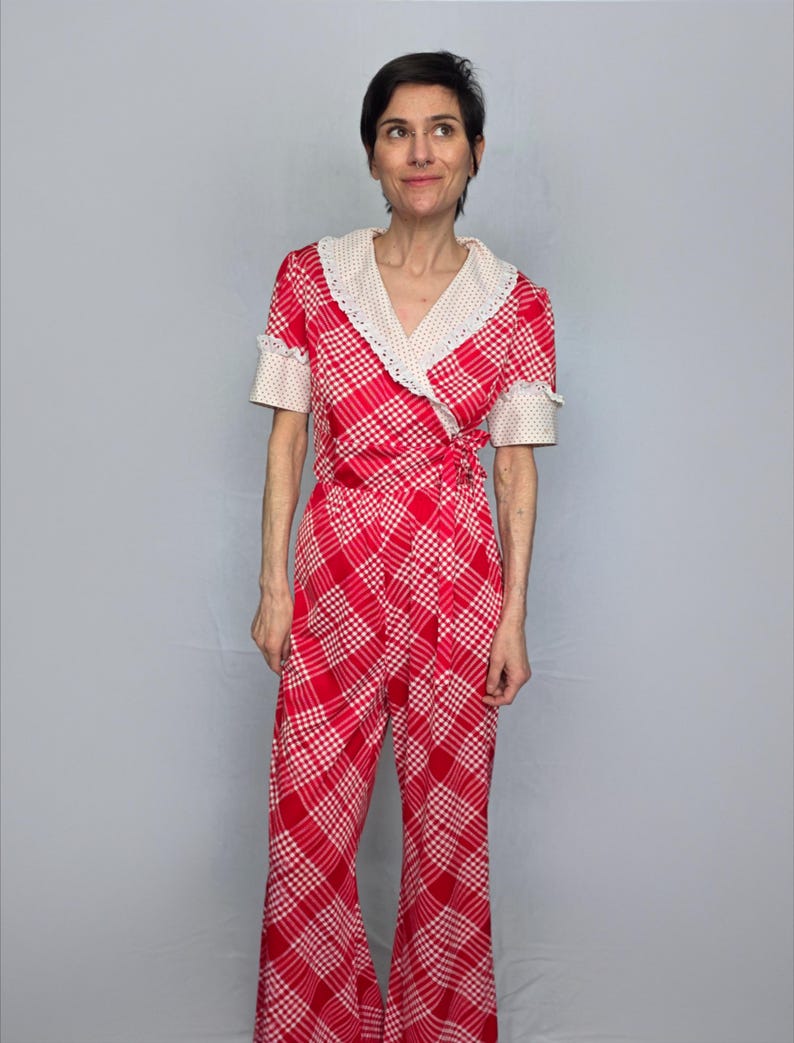 A woman wearing a red and white checkered jumpsuit with a lace collar and short sleeves stands against a gray background.