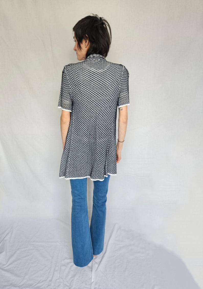 May include: A black and white patterned tunic with short sleeves and a white trim. The tunic is worn over blue jeans.