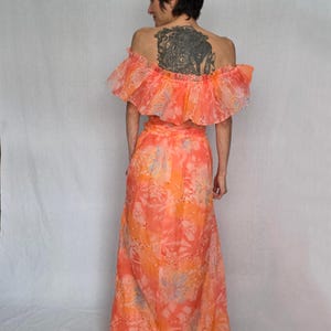 A woman wearing a vibrant, floral-patterned dress with off-the-shoulder sleeves stands against a plain white background.