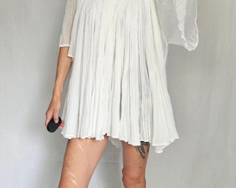 May include: White, short-sleeved dress with a V-neck and pleated skirt. The dress has a crochet neckline and flared sleeves. The person is holding a black object.