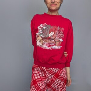 A woman wearing a red sweater with a Christmas-themed design and red plaid pants stands against a gray background.
