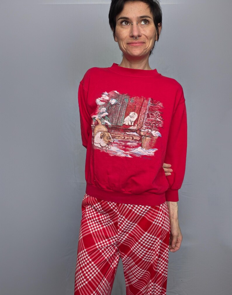 A woman wearing a red sweater with a Christmas-themed design and red plaid pants stands against a gray background.