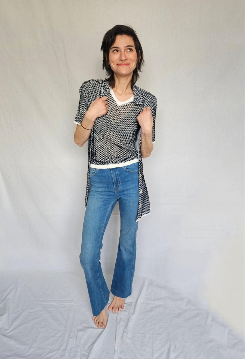 May include: A woman wearing a white and black patterned shirt over a white mesh top and blue jeans.