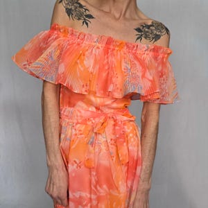 A woman wearing a vibrant, floral-patterned dress with ruffled details and tattoos on her shoulders.