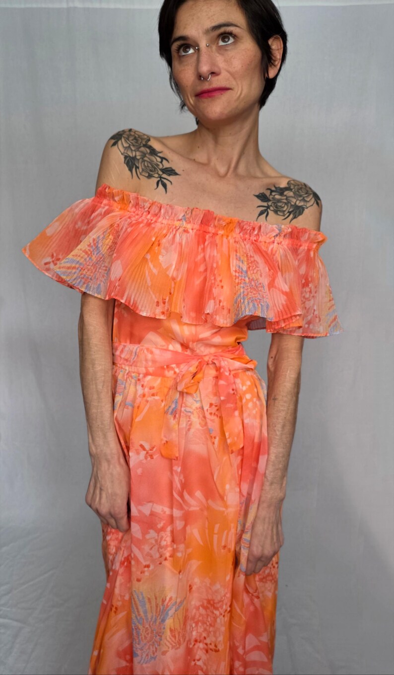 A woman wearing a vibrant, floral-patterned dress with ruffled details and tattoos on her shoulders.